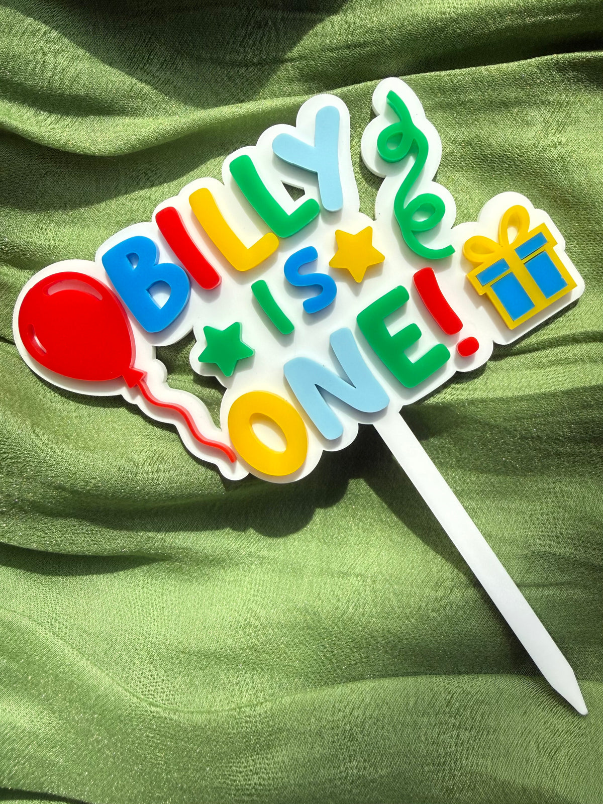 Colourful first birthday cake topper reading Billy is one, with a red balloon, stars and a blue and yellow present.